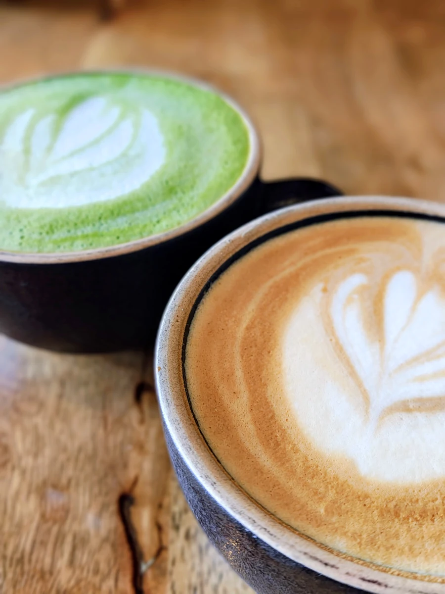 Ahşap masada yan yana duran, süt köpüğü desenli kahve ve matcha fincanları.
