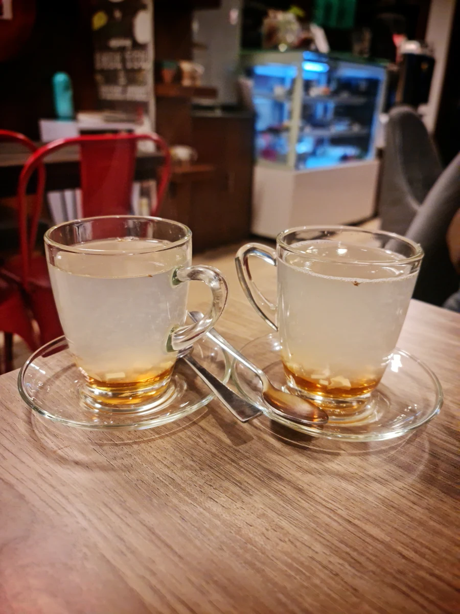 Ahşap masada, cam tabaklar üzerinde açık renkli içecek dolu iki kulplu bardak ve kaşıklar.